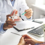 A dentist in a white coat is holding a dental model while pointing to it with a pen, explaining something to a patient. The patient is partially visible, sitting across the desk with hands folded. On the desk, there is a keyboard, a clipboard, and a small potted plant. The setting suggests a dental consultation in a professional office.