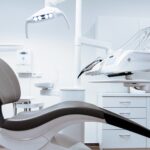 A modern dental office with a sleek, ergonomic dentist chair in dark gray and white. Overhead, there is a bright dental light fixture, and to the side, a set of dental tools are mounted on an adjustable arm. The room is clean and well-organized with white cabinetry and drawers, creating a professional and sterile environment.