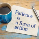A blue coffee cup filled with black coffee sits on a wooden table with a blue distressed finish. Next to the cup, a metal pen rests on a napkin with the handwritten message, "Patience is a form of action." The scene conveys a moment of reflection and motivation, enhanced by the rustic background and the simple yet profound message.