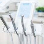 A close-up of various dental tools and instruments arranged in a dental clinic. The equipment includes a scaler, drill, air/water syringe, and other dental handpieces, all neatly placed in their respective holders. The background is slightly blurred, showing a clean and modern dental office setting with a window and a potted plant. The image emphasizes the professional and hygienic environment of the dental care facility.