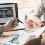Two people are sitting at a desk, discussing dental care. One person, likely the dentist, is holding a pen and pointing to a document, while the other person listens attentively with hands clasped. On the desk, there is a laptop displaying a dental X-ray, a model of teeth, and a small potted plant. The setting suggests a dental consultation, focusing on patient education and treatment planning.