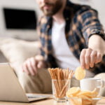 A man in a casual plaid shirt is sitting on a couch with a laptop in front of him. He is reaching for a potato chip from a bowl on the table. The table also has a glass filled with pretzel sticks and a white mug. The background is blurred, showing a cozy living room with a soft focus on the man, emphasizing a relaxed, casual setting.