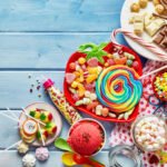 A colorful assortment of candies and sweets spread out on a light blue wooden table. The arrangement includes a large rainbow swirl lollipop, various gummy candies, candy canes, chocolate bars, and cookies. There are also lollipops, sprinkles, and a bowl of bright red ice cream with rainbow sprinkles. A polka dot napkin and vibrant plastic spoons add to the festive and cheerful presentation. The image conveys a fun and indulgent treat selection.