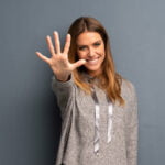 A young woman with long brown hair is smiling and extending her hand towards the camera, showing all five fingers. She is wearing a gray hoodie with ribbon drawstrings, standing against a solid gray background. The gesture and expression convey a friendly and welcoming attitude.
