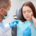 A woman sitting in a dental chair is touching her cheek with a pained expression, indicating dental discomfort. A dentist wearing a mask and blue gloves holds a dental mirror and speaks to her. The woman is wearing a striped shirt and a blue dental bib. The background shows a clinical setting, emphasizing a visit to the dentist for tooth pain or a dental check-up.
