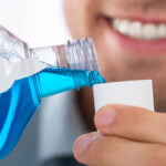 A close-up of a man pouring blue mouthwash into a white cap. The man is smiling, showing his white teeth. The focus is on the mouthwash bottle and cap, with the man's mouth slightly blurred in the background. The image emphasizes oral hygiene and the use of mouthwash as part of a dental care routine.