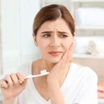 A woman is holding a toothbrush with orange and white bristles in her right hand while touching her cheek with her left hand. She has a pained expression, indicating discomfort or a toothache. She is wearing a white shirt, and the background shows a bright, clean bathroom with shelves of neatly folded towels. The image highlights dental pain or issues while brushing teeth.