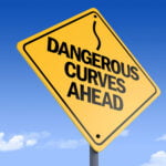 A yellow diamond-shaped road sign with black lettering reads "DANGEROUS CURVES AHEAD" and includes a wavy arrow symbol above the text. The sign is set against a clear blue sky with a few white clouds, indicating a sunny day. The image serves as a warning to drivers about upcoming winding or hazardous road conditions.