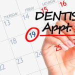 A close-up of a hand marking a calendar with a black marker. The date "19" is circled in red, and the words "DENTIST Appt.!" are written across the calendar on the 19th, highlighted with a red underline. The days of the week are labeled, and the numbers are in a mix of blue and red, with the background of the calendar being white. The image emphasizes the importance of remembering a dental appointment.