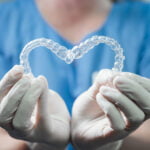 A dental professional wearing blue scrubs and white gloves is holding two clear aligners, forming them into a heart shape. The close-up focuses on the aligners and hands, emphasizing the dental care theme. The background is blurred, keeping attention on the transparent aligners and the gloves. The scene highlights orthodontic treatment and care.