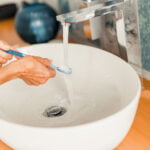 A close-up of hands rinsing a blue toothbrush under running water in a white, modern sink. The bathroom features a chrome faucet and a wooden countertop. In the background, there is a blue decorative vase and a tiled backsplash with blue and white patterns. The scene highlights a daily oral hygiene routine.