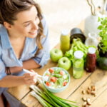 A woman with long brown hair, wearing a blue and white striped shirt, is smiling while sitting at a wooden table filled with fresh vegetables, fruits, and jars of juice. She is holding a fork and eating a salad with cherry tomatoes, lettuce, and cucumbers. The table also features green apples, green onions, an avocado, and a variety of leafy greens, creating a vibrant and healthy dining scene. The background is bright and airy, enhancing the fresh and wholesome atmosphere.