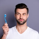 A man with dark hair and a beard is holding a blue toothbrush, looking at it with a slightly raised eyebrow and a thoughtful expression. He is wearing a white t-shirt, and the background is a plain, muted gray, emphasizing his focus on the toothbrush.