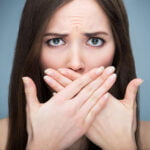 A young woman with long brown hair covering her mouth with both hands. Her blue eyes are wide open, and her eyebrows are furrowed, conveying a sense of shock, concern, or embarrassment due to bad breath. The background is a plain, muted blue, keeping the focus on her facial expression.