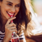 A woman with a radiant smile, wearing a striped top, sipping a drink through a red and white striped straw. The sunlit setting gives a warm, inviting atmosphere, capturing a moment of leisure and enjoyment.