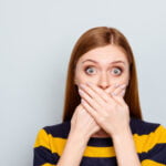 A woman with wide eyes covers her mouth with both hands, wearing a yellow and navy striped sweater. Her expression suggests shock or surprise, set against a plain light gray background.