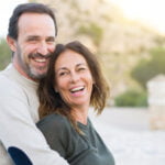 A joyful middle-aged couple embracing and smiling outdoors, with the man behind the woman, both looking into the camera. Their relaxed, genuine smiles and casual attire suggest a moment of happiness and comfort in each other's company. The background is softly blurred, highlighting their connection.