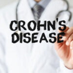 A medical professional in a white coat writing 'CROHN'S DISEASE' on a transparent surface with a black marker, symbolizing education or awareness about the condition.