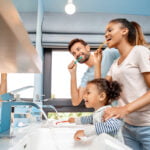 A happy family engaged in their dental hygiene routine; a man and woman brushing their teeth with a toddler, all smiling, in front of a bathroom mirror.