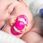 A peaceful sleeping baby with a pink pacifier in their mouth, nestled in soft white bedding. The baby's delicate eyelashes are highlighted in a close-up, and a hint of a cozy blue and purple knit blanket adds a touch of warmth to the tranquil scene.