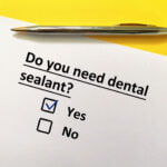 A piece of paper with the question "Do you need dental sealant?" printed on it, with a checkbox next to "Yes" marked with a blue checkmark and an unmarked checkbox next to "No." A silver pen lies above the paper on a vibrant yellow background, suggesting the completion of a dental questionnaire.
