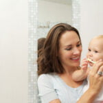 A smiling woman holding a toddler is teaching the child how to brush teeth, using a small yellow toothbrush. They are in a bathroom with a tiled wall and a mirror reflecting part of the scene, emphasizing a nurturing, domestic moment focused on dental care.