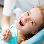A young girl reclining in a dental chair holds a dental mirror, showing an open-mouthed smile with missing teeth, indicating a child's dental examination in progress.