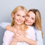 A heartwarming portrait of two women, likely mother and daughter, with joyful expressions and blonde hair, hugging each other. The older woman is wearing a pink cardigan over a white shirt, while the younger one is in a light blue sweater, both against a soft grey background that enhances their bright smiles.