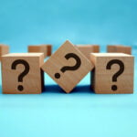 Three wooden blocks with question marks facing forward, with a blurred array of similar blocks in the background, all set against a bright blue surface, symbolizing problem-solving, decision-making, or the search for answers.