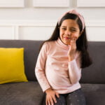 A young girl sitting on a couch holding her cheek, showing a discomforted expression that suggests toothache or dental pain, with bright yellow pillows adding a contrasting color to the scene.