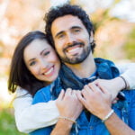 A happy couple embracing outdoors with a blurred nature background. The woman, with long dark hair, is hugging the man from behind and smiling widely. The man, with curly hair and a beard, is holding her hands around his chest, sharing a joyful moment together.