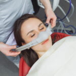 A woman reclined in a dental chair with her eyes closed, receiving nitrous oxide sedation through a nasal mask held by a dental professional. The patient appears relaxed, and the environment suggests a dental procedure is about to take place.