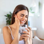 Woman with long brown hair, wearing a white tank top sits in her living room while holding a cold drink in her left hand. Her right hand is holding the side of her face while she winces in pain. She sits on a white couch. Behind her is a white dining table, a desk, a green plant and a window letting in natural light. Bruxism, teeth grinding and teeth clenching is common and painful. We can help!