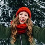 A woman with long reddish blond, curly hair stands with arms outstretched, wearing a forest green winder coat, red scarf and red hat. She smiles big while sticking out her tongue in a playful manner. Behind her is a tree covered in snow.