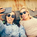 Two young women lying on a wooden surface, playfully sticking their tongues out as they take a selfie. They each have a tongue piercing. Both wear sunglasses, with one in a denim shirt and an anchor tattoo on her wrist, and the other in a beige top holding a skateboard. The mood is casual and fun.