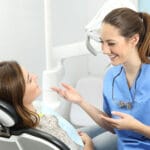 A female dental hygienist in blue scrubs is smiling and talking with a female patient who is reclining in a dental chair. The patient is wearing a disposable dental bib and appears relaxed. Dental equipment and a bright, clean clinic setting are visible in the background.