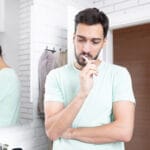 A man in a light green shirt stands in a modern bathroom, brushing his teeth while appearing thoughtful. He has his arms crossed and gazes downward, with a mirror reflecting his side profile in the background. The bathroom features white brick tiles, a towel rack, and a doorway leading to a wooden interior.