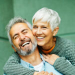 A happy older couple with light skin is embracing and laughing together. The woman, with short white hair, is wearing a textured green sweater and is hugging the man from behind, resting her head against his. The man, with salt-and-pepper hair and a beard, is wearing a light blue button-up shirt and smiling with his eyes closed. The background is a muted green wall.