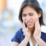 Woman with long dark hair and a navy collared shirt touching both sides of her jaw with a pained expression, indicating toothache or jaw pain.