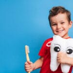 Smiling young boy in a red shirt holding a yellow toothbrush and a plush toy shaped like a tooth with big cartoon eyes, standing against a bright blue background.