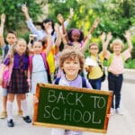 Smiling boy holding a chalkboard with “Back to School” while classmates with backpacks raise their hands outdoors.