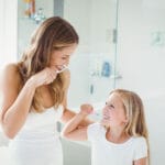 Woman and young girl smiling while brushing their teeth together in a bright bathroom.